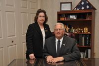 A man in a suit sits at a desk with a woman standing beside him in an office with a bookshelf and a flag.