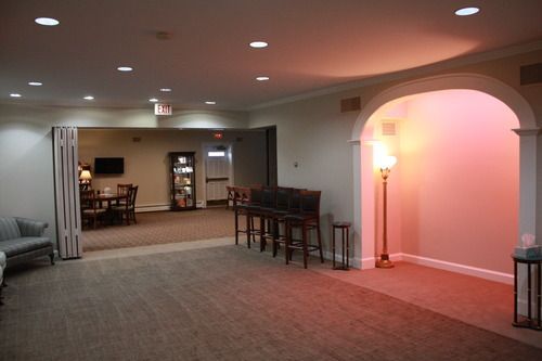 A spacious room with beige carpet, a seating area, a bar, and an arched alcove illuminated by a soft, pink light.