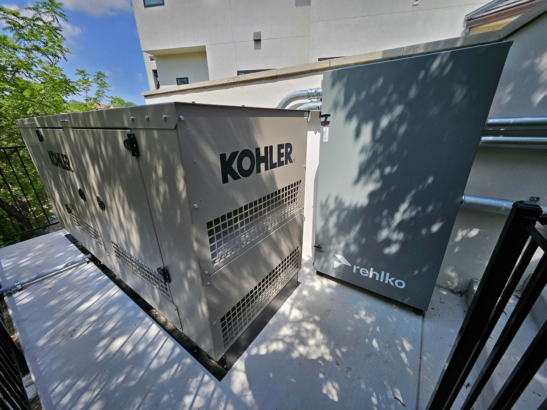 Two outdoor generators beside a building, one white Kohler unit and one dark blue unit in sunlight.