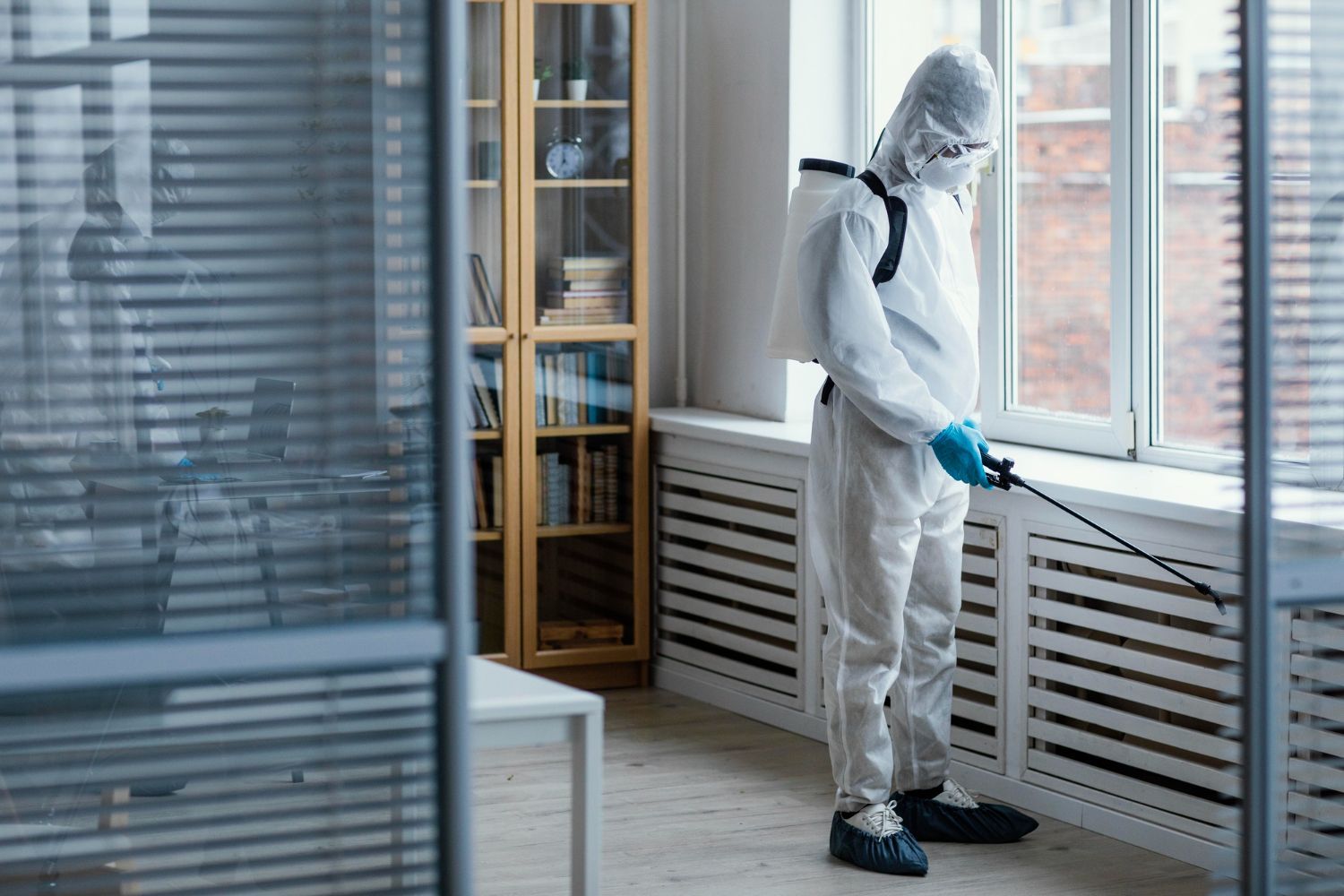 Worker in white protective suit disinfecting an office with a sprayer near windows