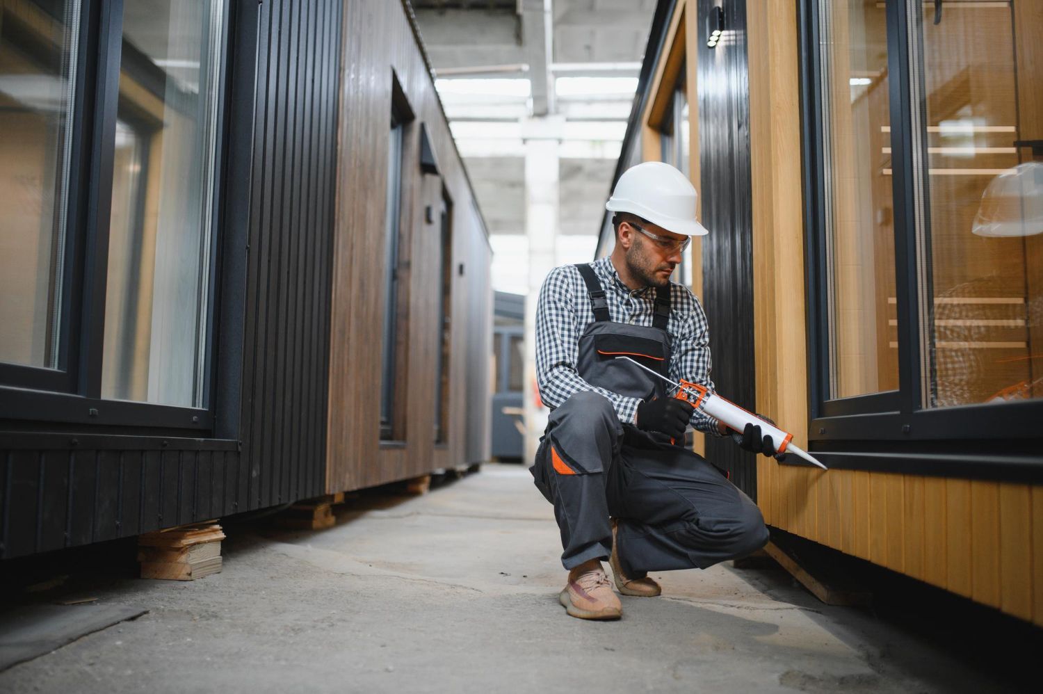 Worker in hard hat kneeling between shipping containers, using a power drill in a warehouse aisle.
