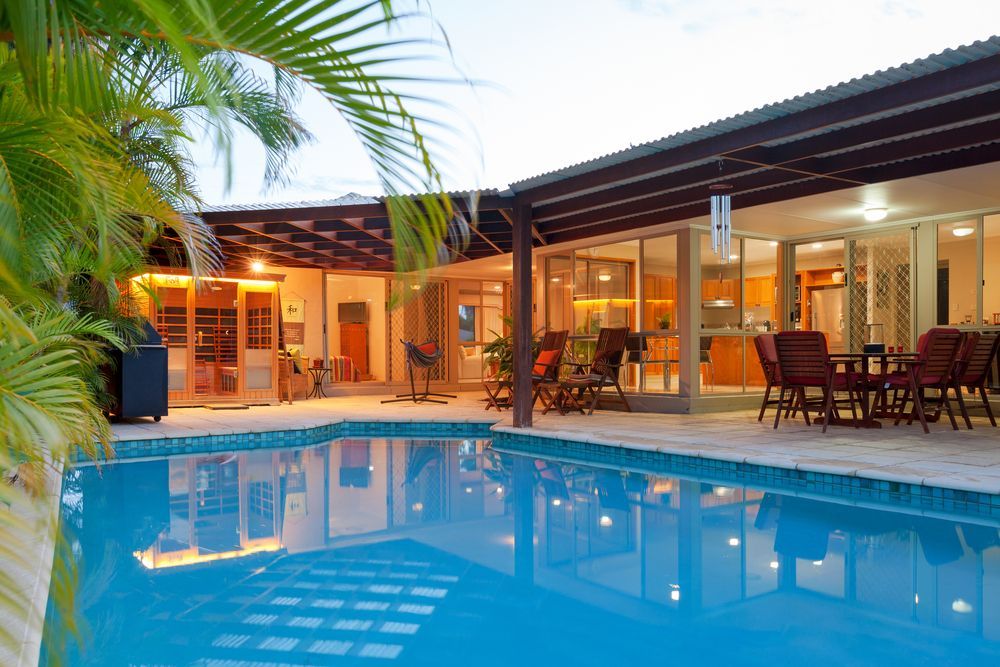 Modern backyard view of a house at sunset with large glass doors, wooden patio furniture, a covered outdoor area, and a swimming pool in the foreground, surrounded by palm leaves.— Lismore Poolscene in Lismore, NSW