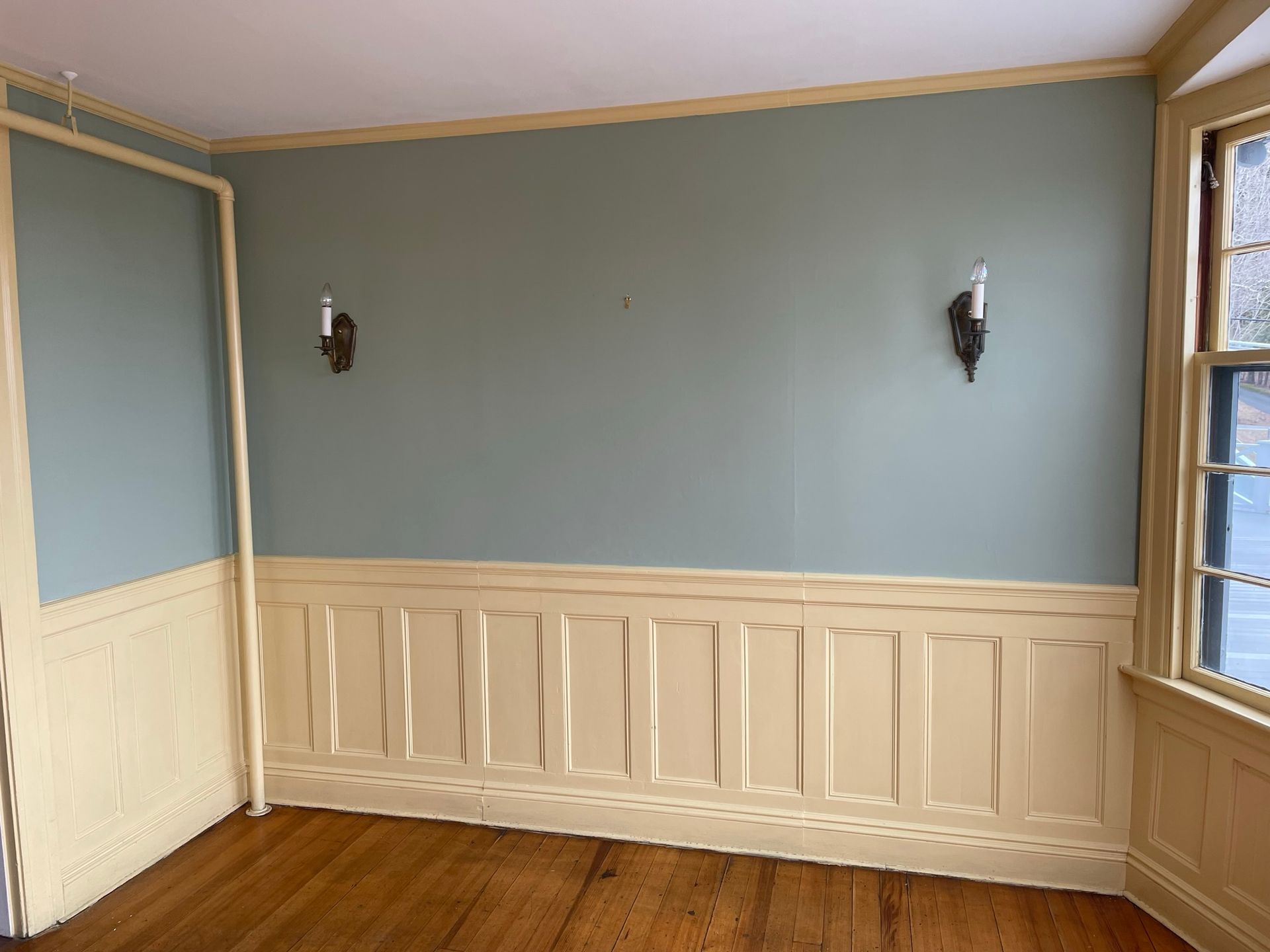 Room with blue-green walls, yellow wainscoting, and sconces. Wooden floor and window visible.