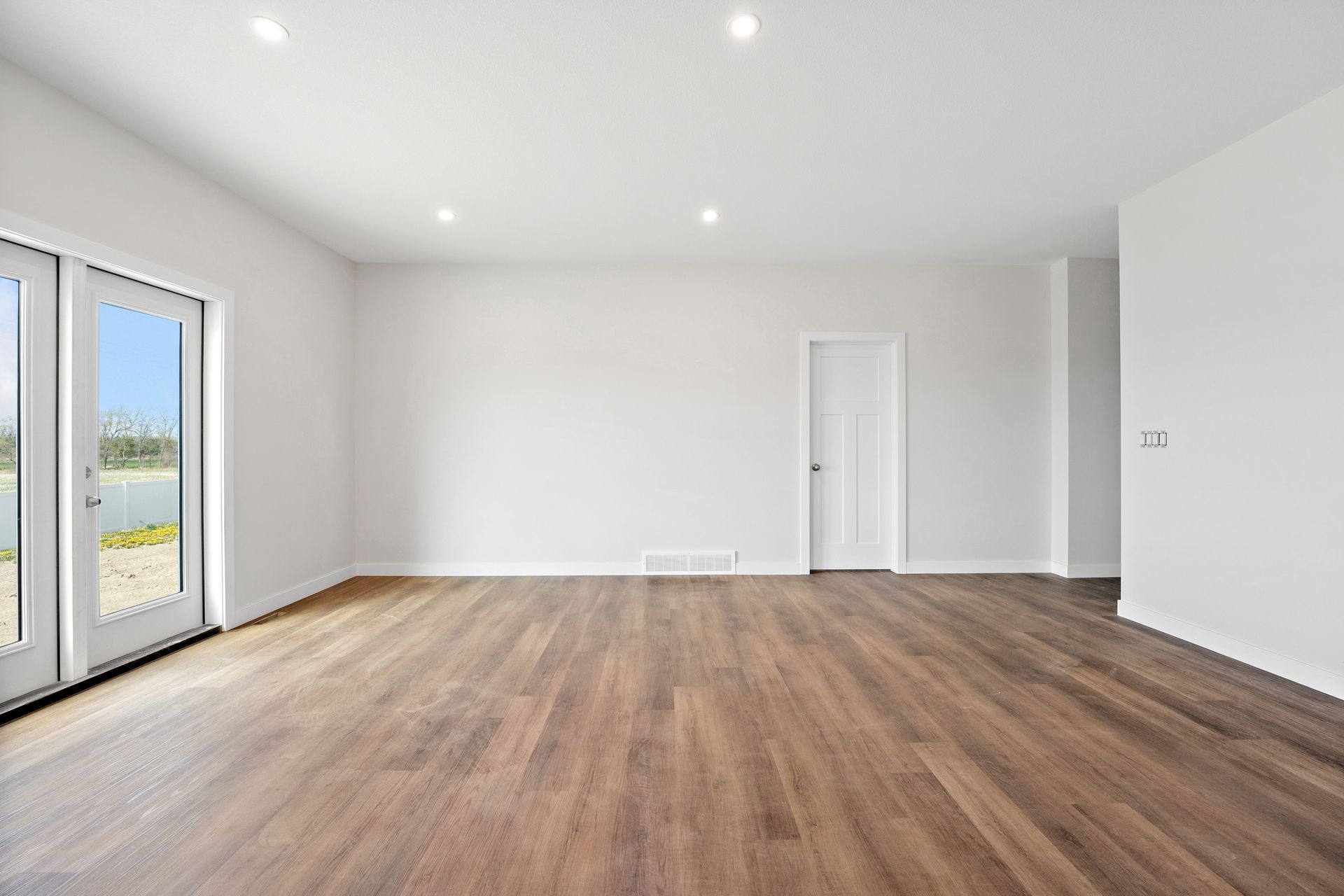 Empty modern living room with white walls, recessed lights, wood flooring, and a glass door to a balcony