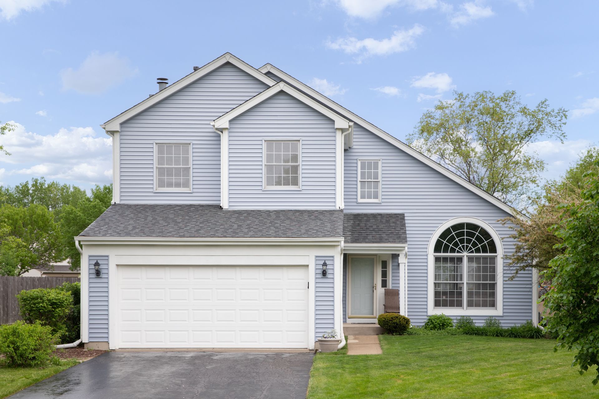 Gray suburban two-story house with a white garage, arched window, and front lawn under a blue sky