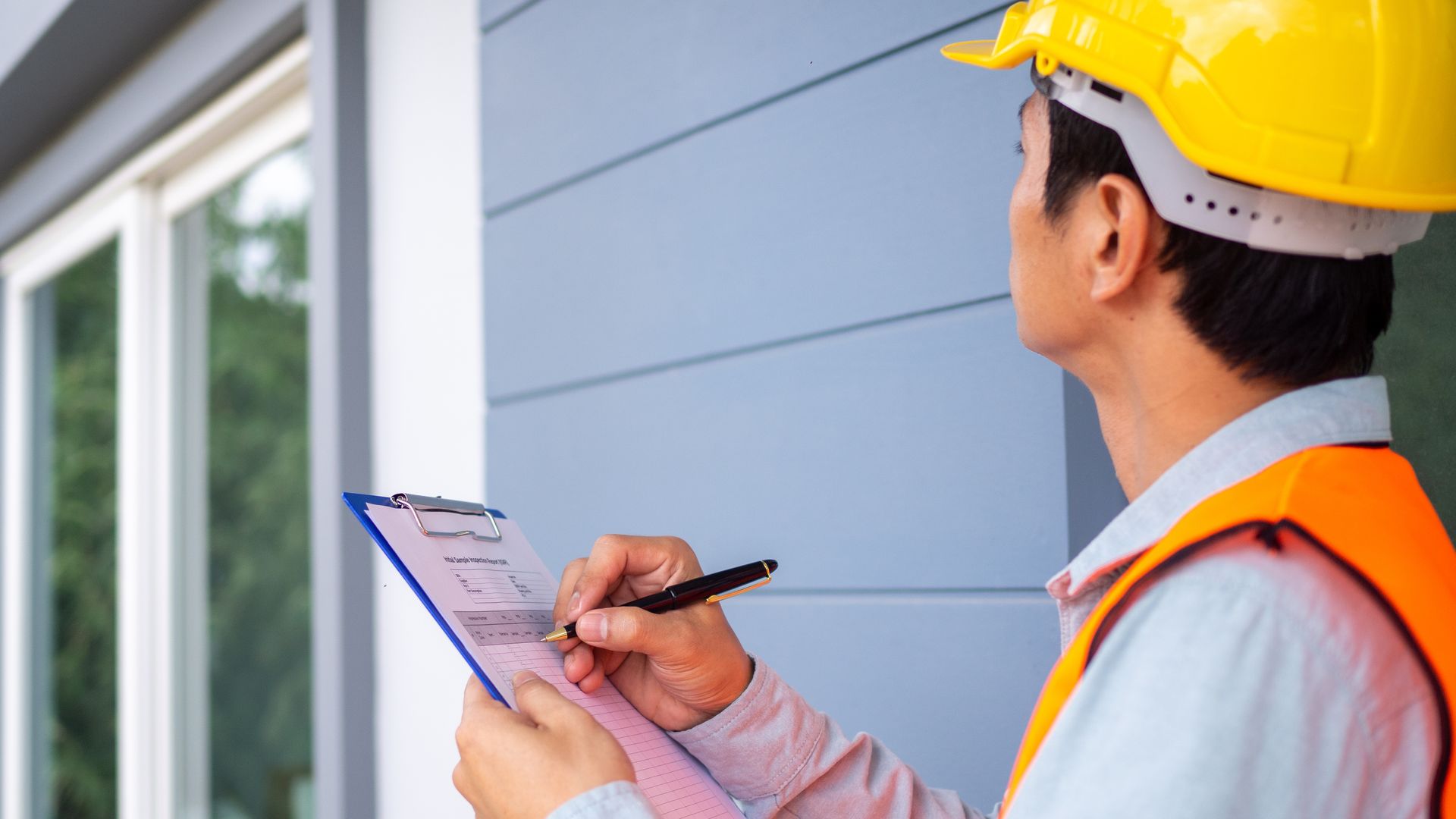 Person in safety vest and hard hat inspecting a building exterior, writing on a clipboard.