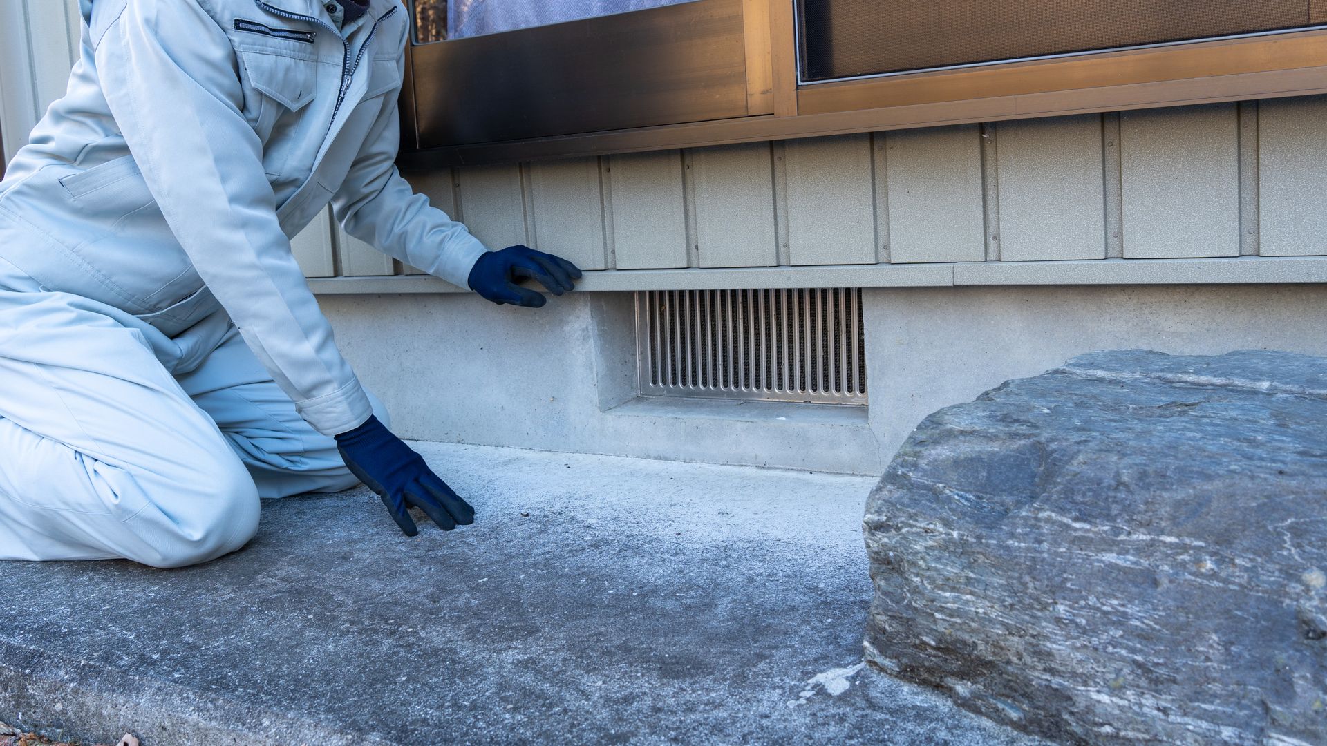 Person kneeling, inspecting a home's foundation. Gray and white building with a ventilation vent, rock in foreground.