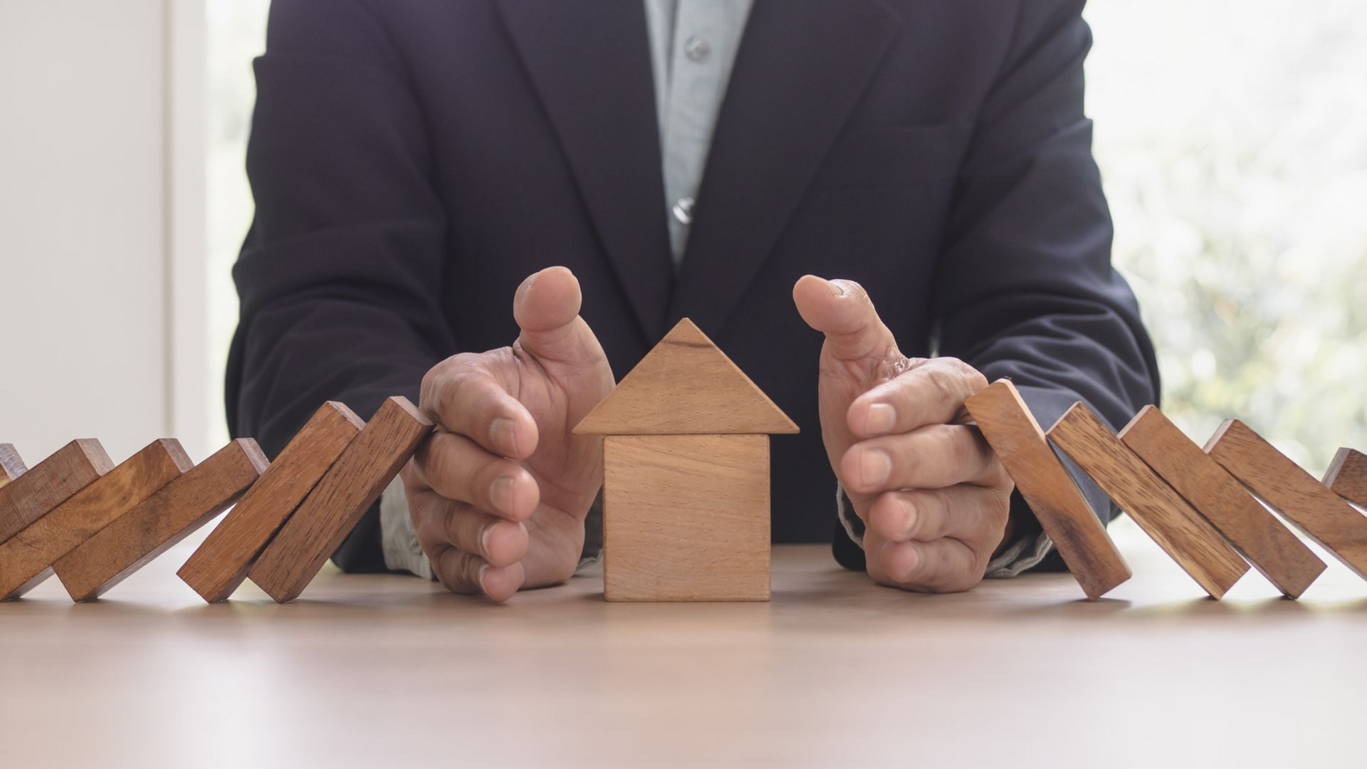 Businessperson shielding a small wooden house from falling dominoes on a table