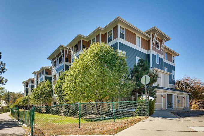 Modern apartment building with blue-gray siding, balconies, and a tree-lined corner entrance