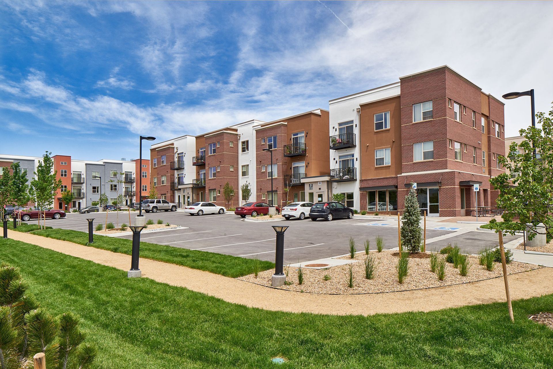 Modern apartment complex with brick buildings, parking lot, and landscaped green lawn under a blue sky