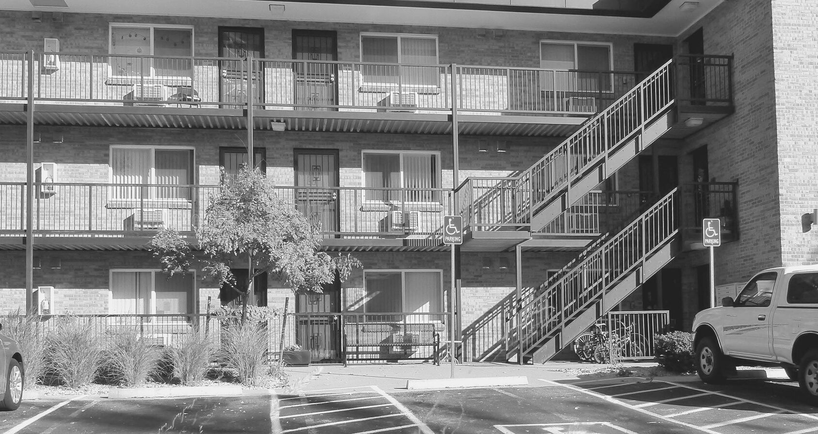 Black-and-white view of a motel-style apartment building with exterior staircases and parked vehicles.
