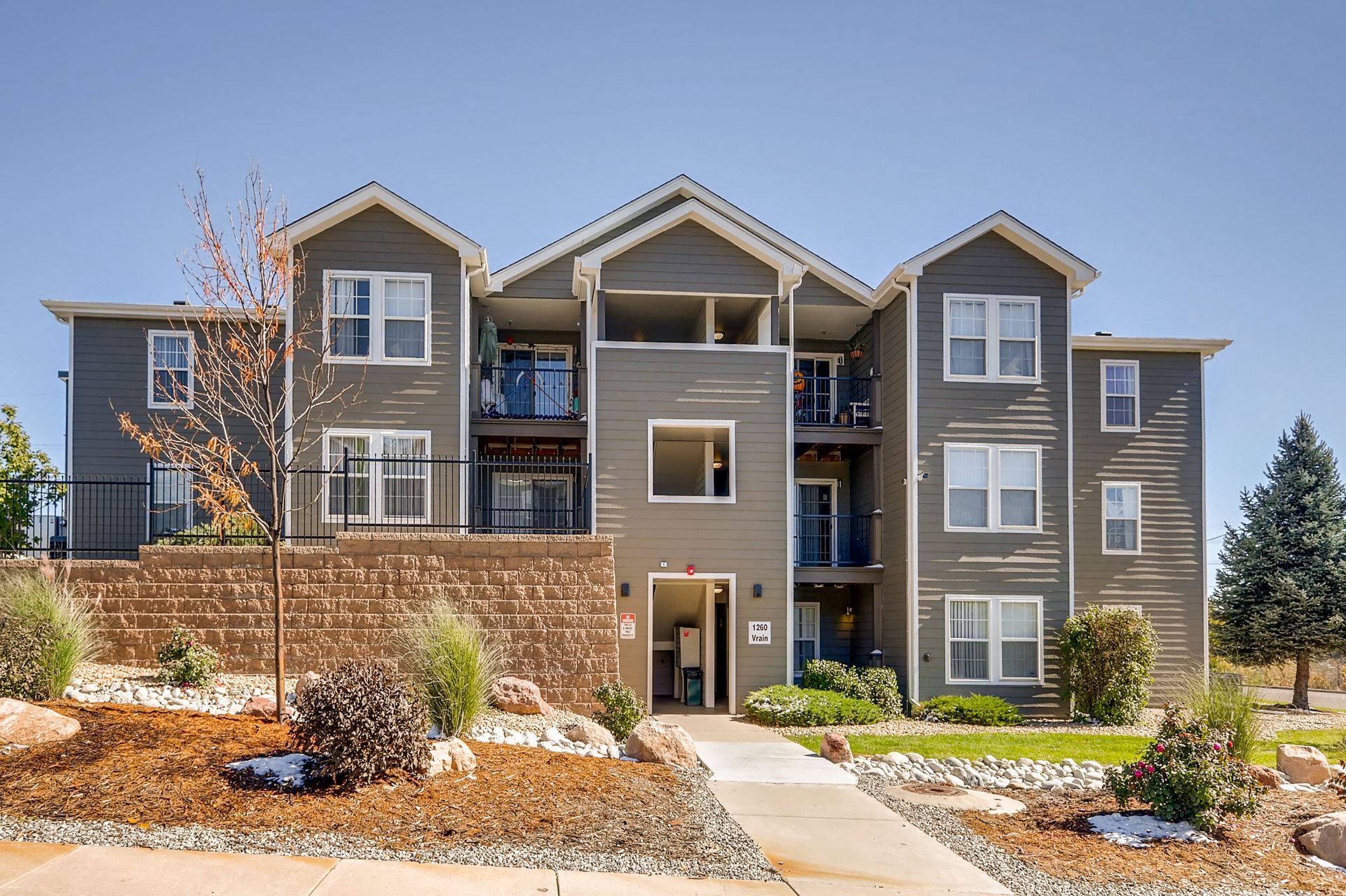Modern three-story apartment building with gray siding, balconies, and landscaped front walkway