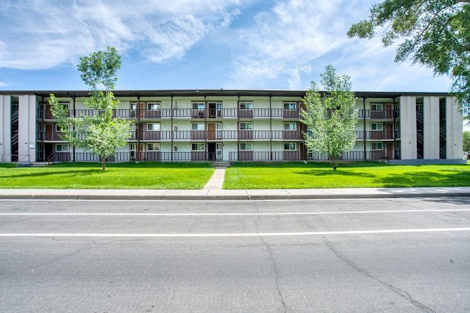 Three-story apartment building with balconies and trees along a roadside under a blue sky