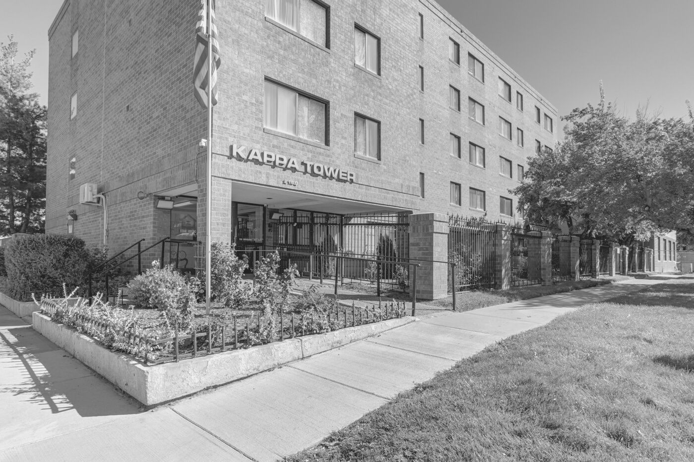 Apartment building entrance with glass awning, sidewalk, and landscaped flower bed on a sunny day
