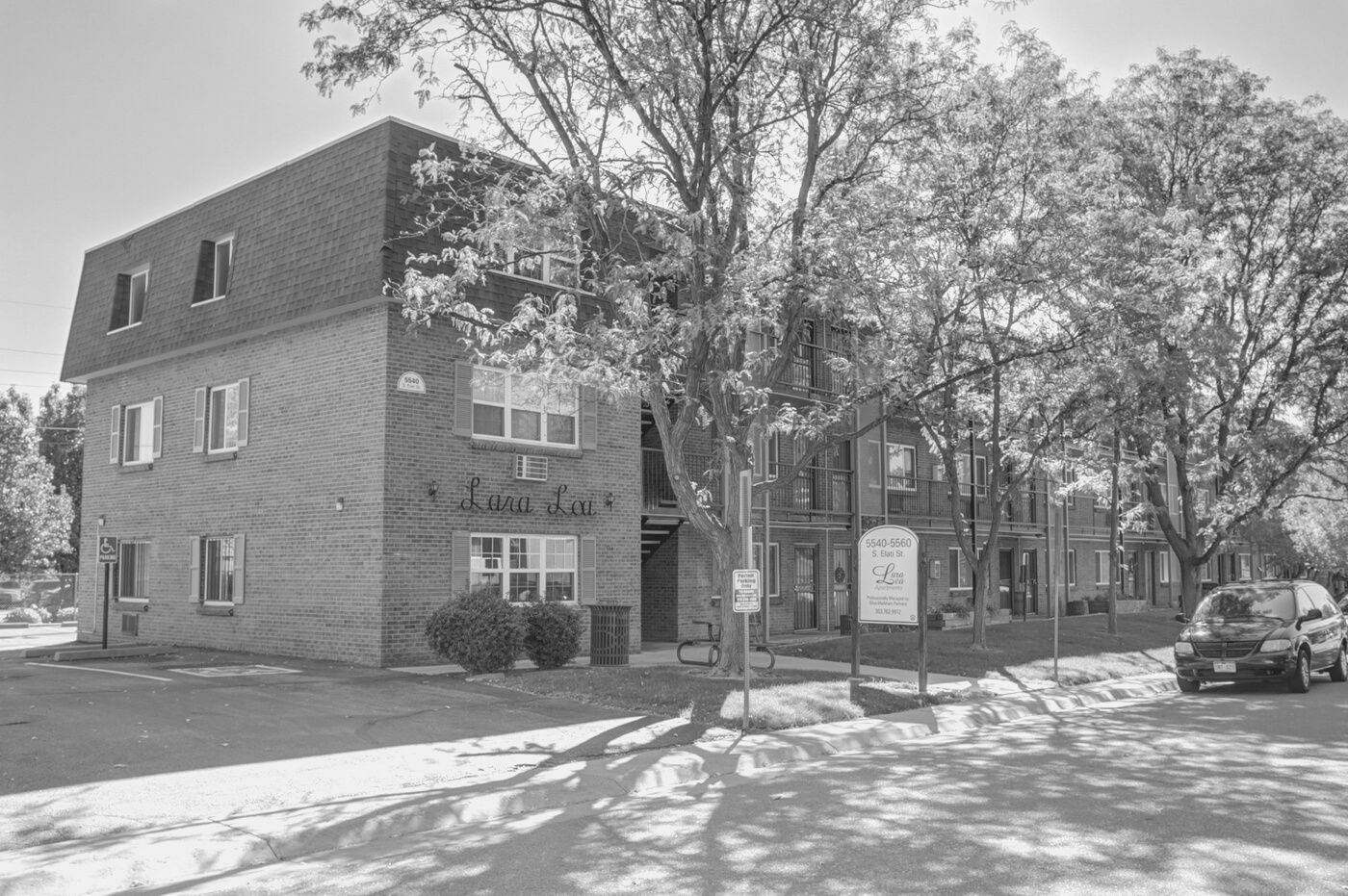Brick apartment building on a tree-lined street with parked cars and a sidewalk sign