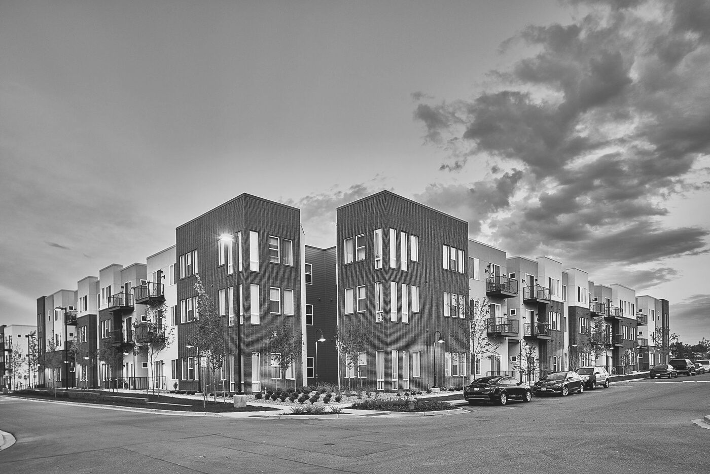 Black-and-white photo of a modern apartment building with parked cars and cloudy sky