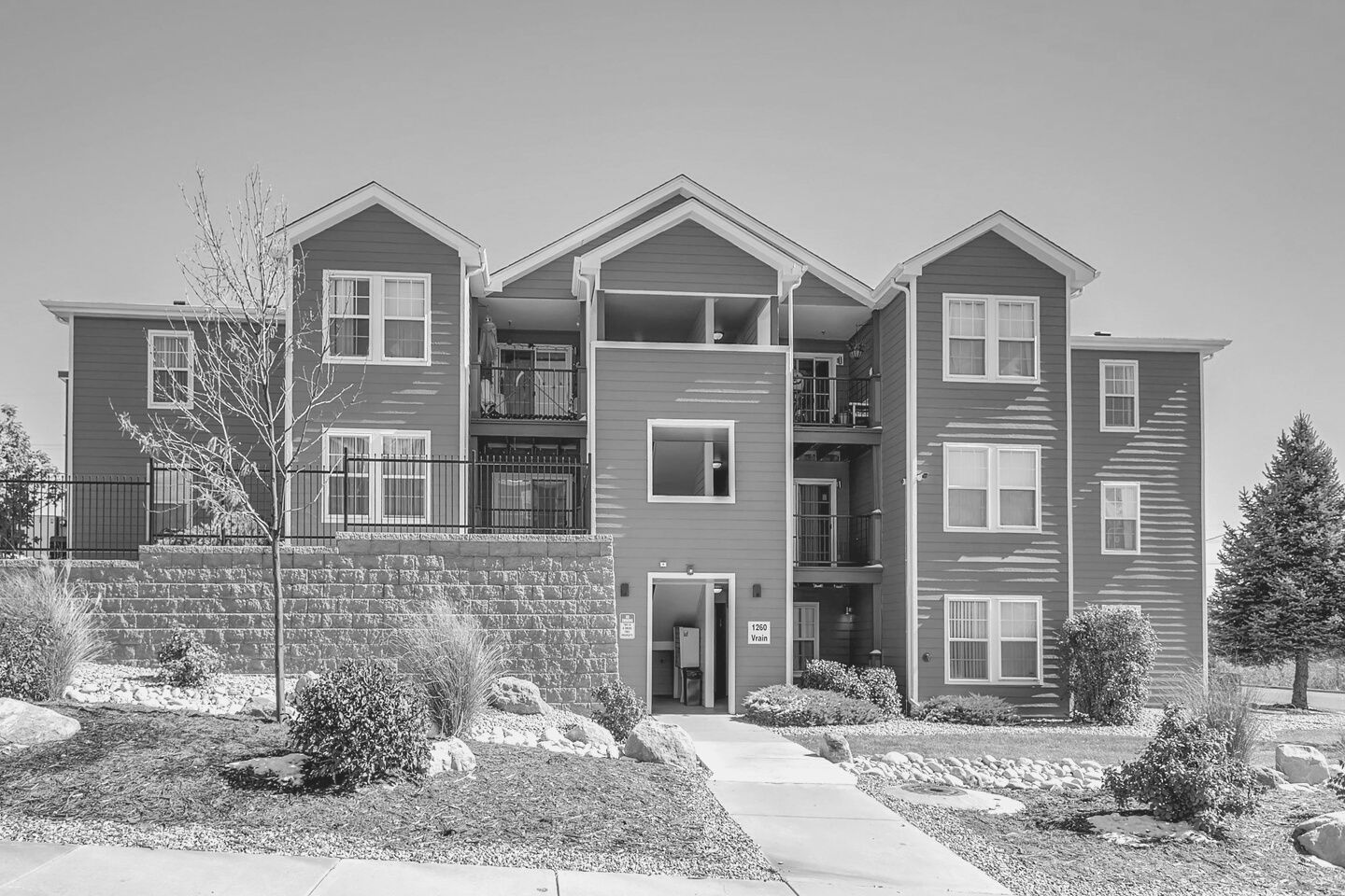 Three-story apartment building with stone facade, balconies, and a landscaped walkway in front.