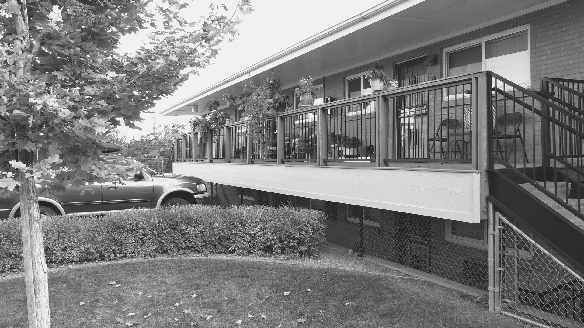 Black-and-white view of a motel walkway with railings, stairs, parked car, and trees alongside the building
