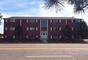 Red brick apartment building with white-trimmed windows behind a road and trees overhead