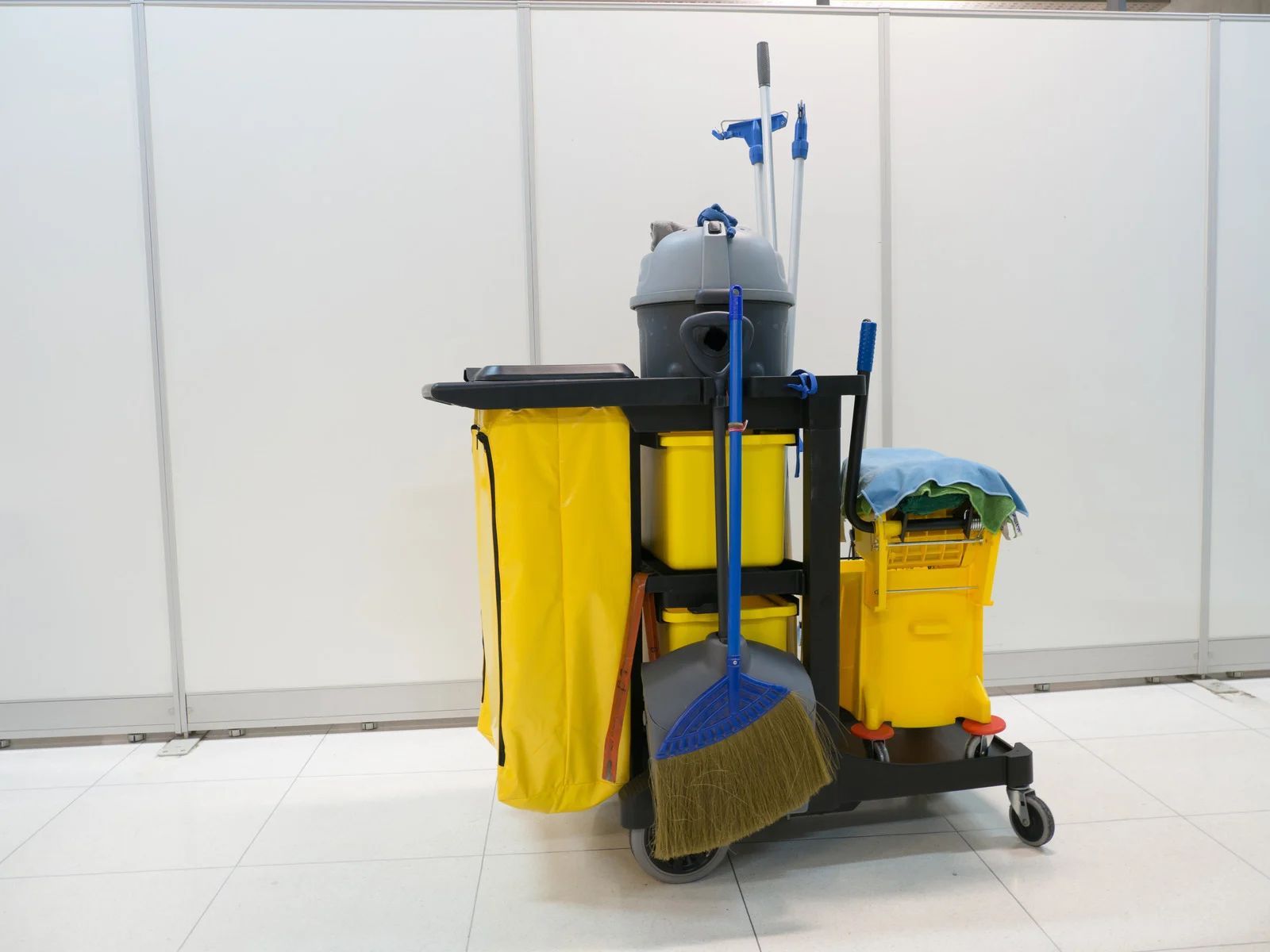 Yellow janitorial cleaning cart with mop, broom, and supplies on a tiled floor.