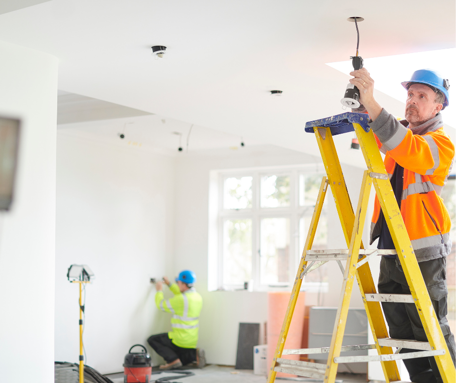 Electrician installing a light fixture in a brightly lit room, wearing safety gear, another electrician working on a wall.
