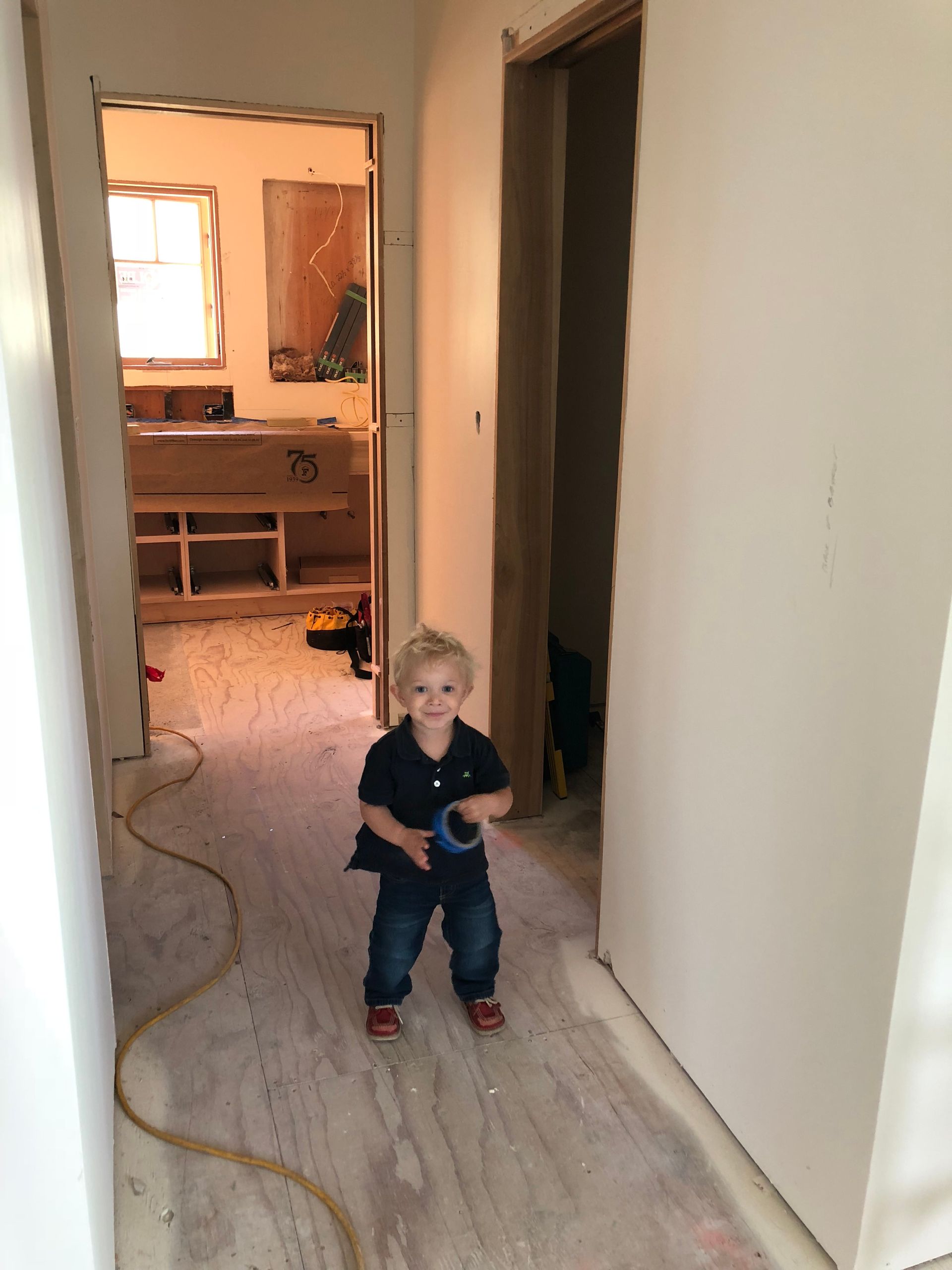 A young child stands in a hallway with construction, holding a blue object. Light-colored walls and flooring are visible.