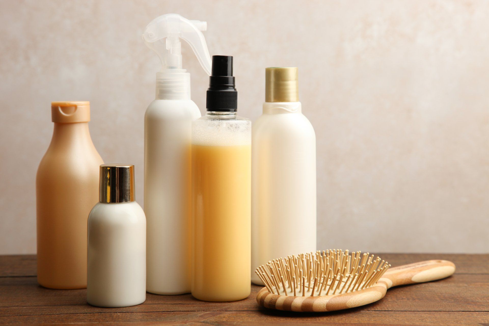 Bottles of hair products and a wooden brush on a wooden surface, neutral background.