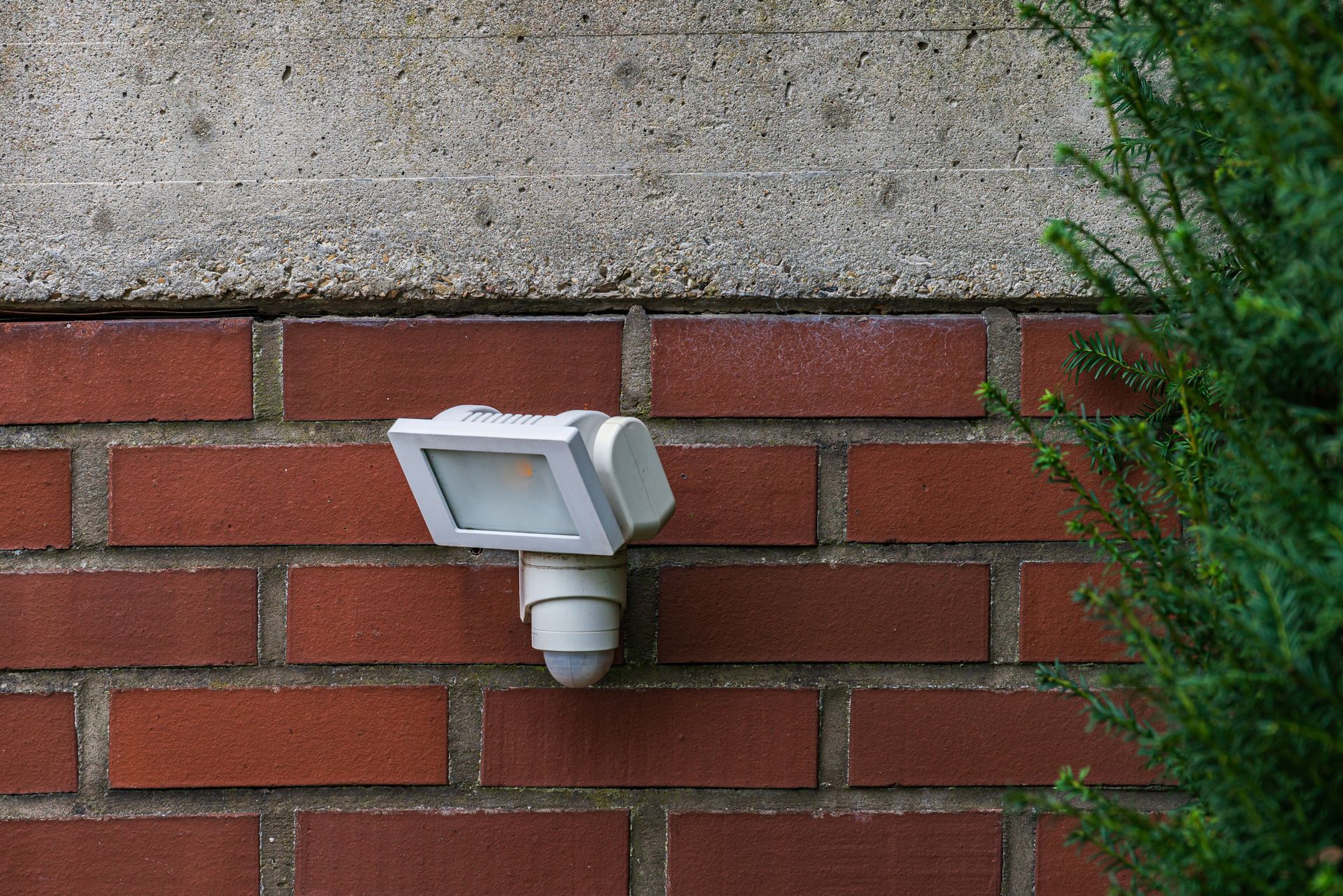 Motion sensor light on a brick wall, next to green bush.