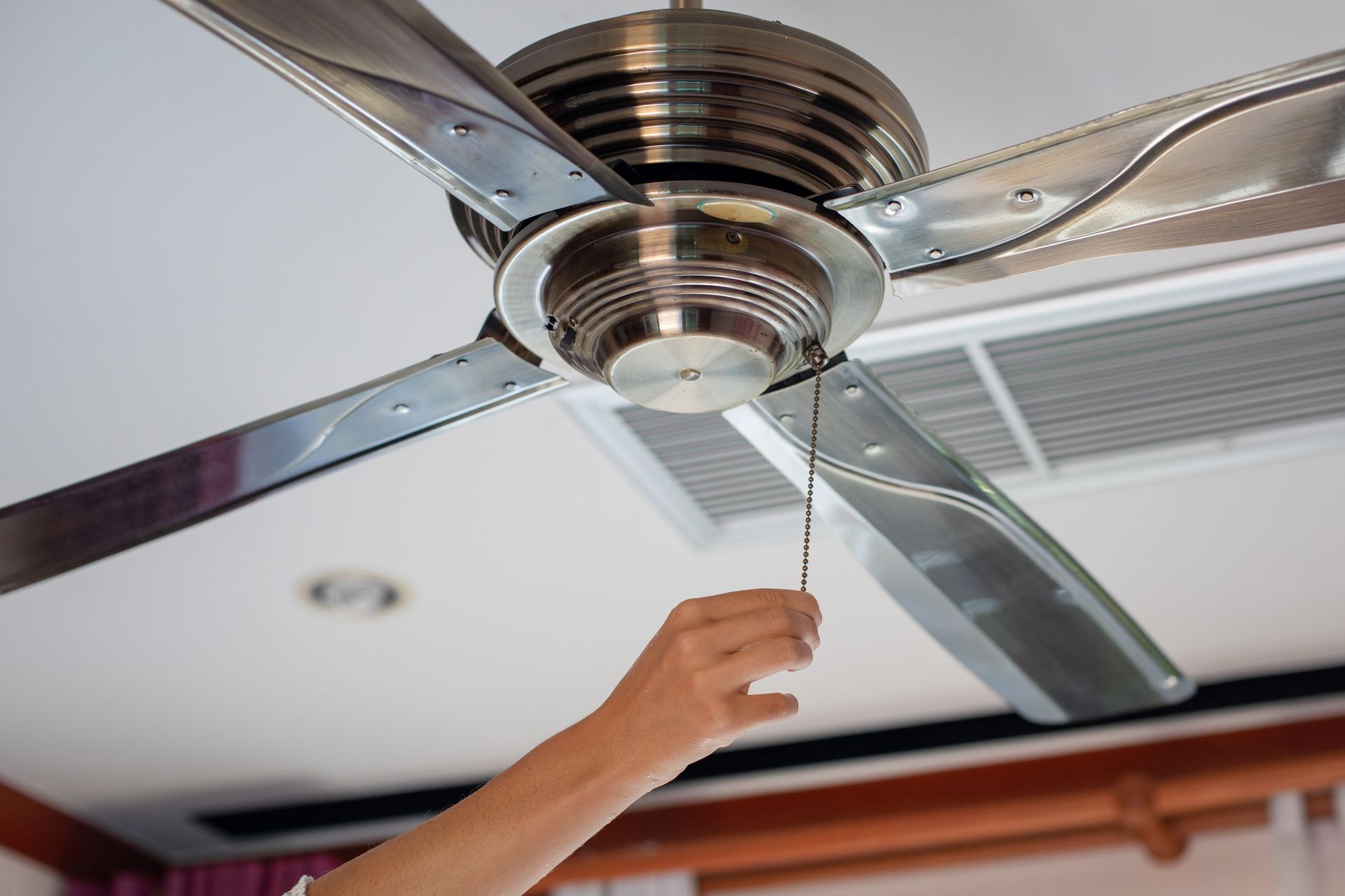 Hand pulling a ceiling fan's chain to turn it on, indoors. Fan has silver blades and a brushed metal finish.