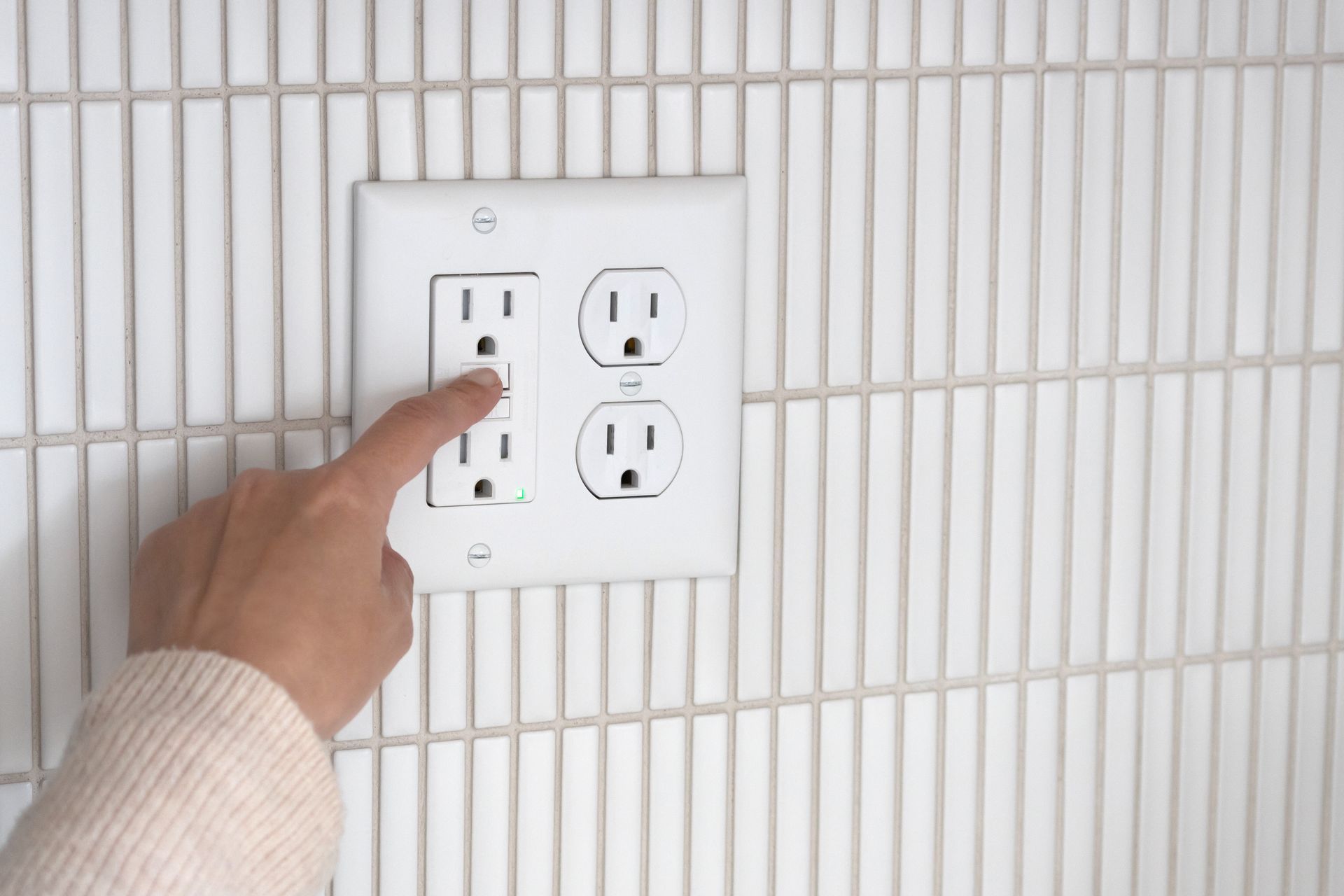 Hand pointing at a vertical outlet on a white tiled wall.