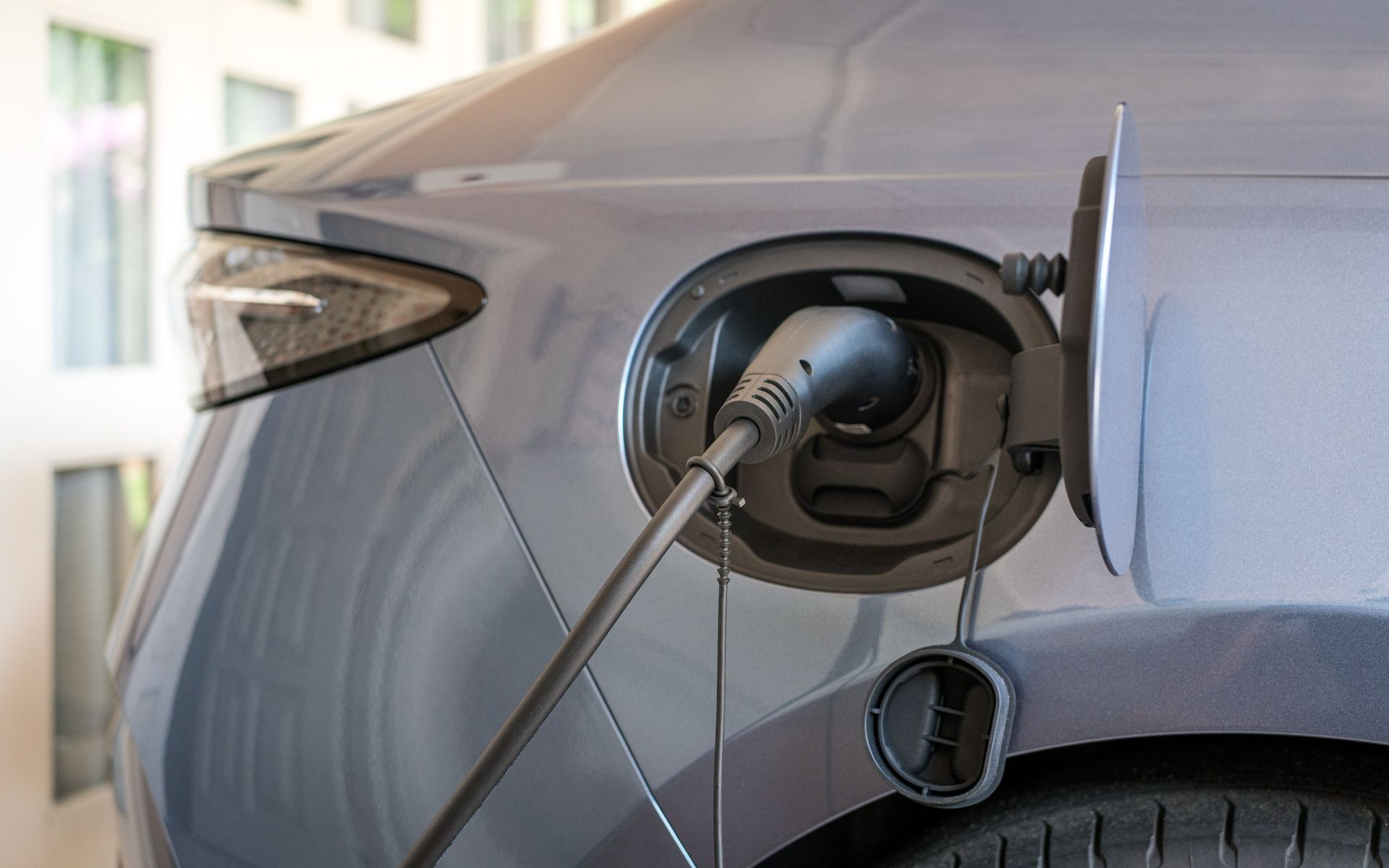 Electric vehicle charging, cord plugged into a car's charging port; gray car, bright background.