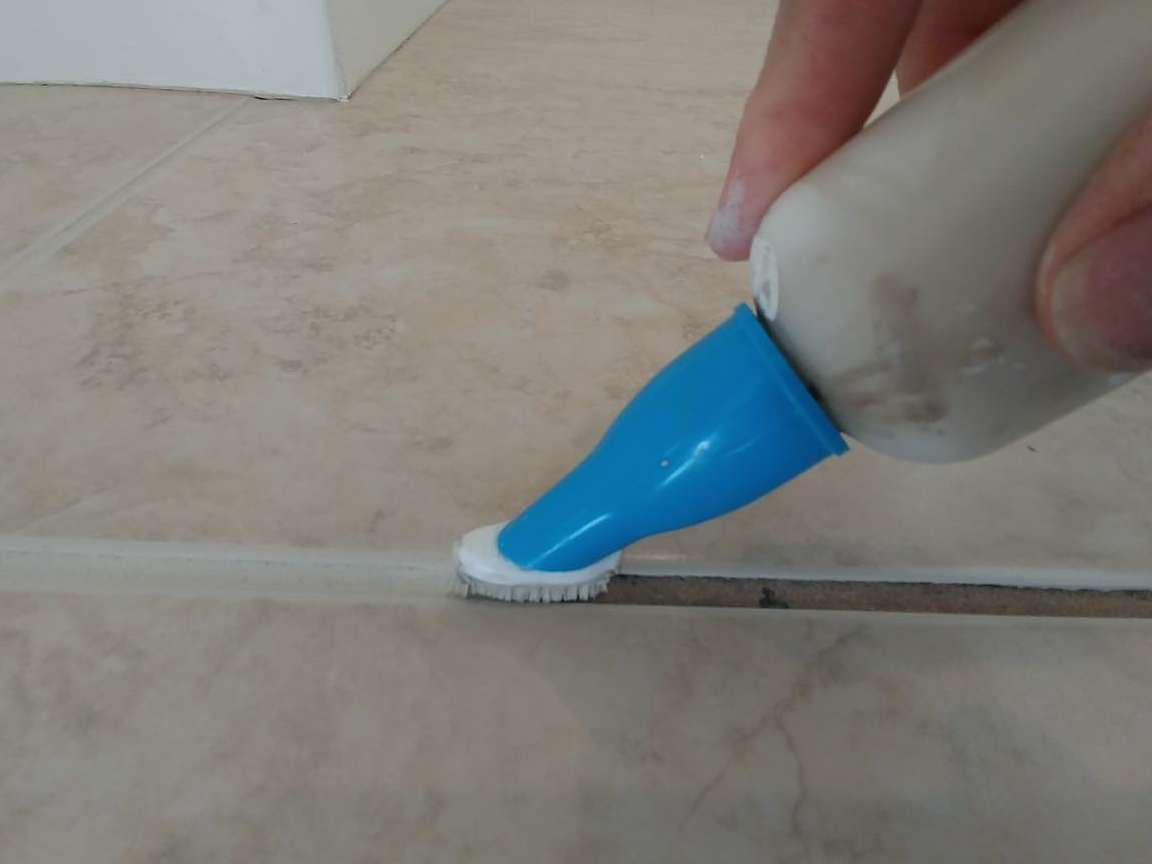 Hand using a cleaning tool with a brush to scrub grout on beige tile.
