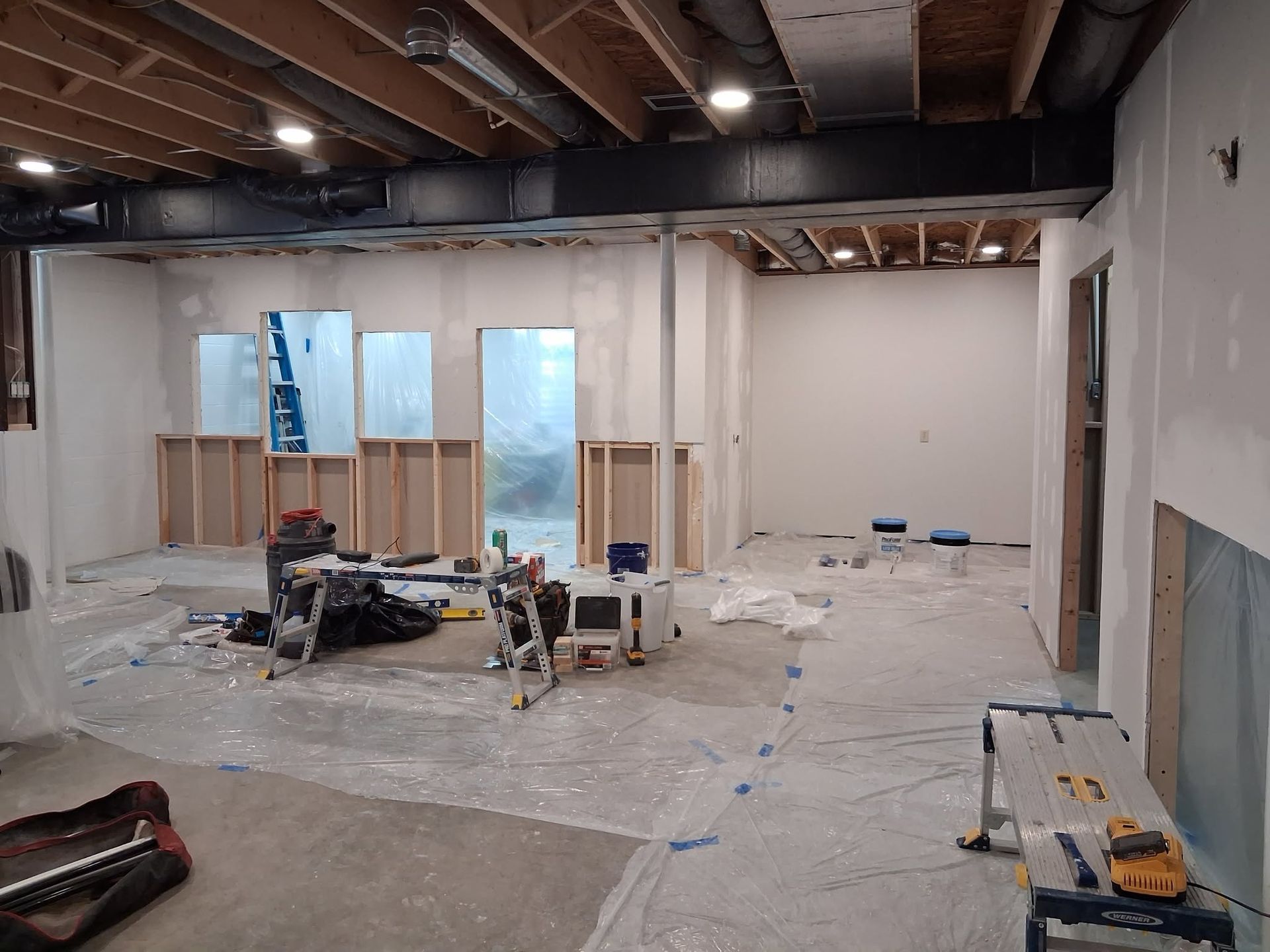 Indoor construction site with framed walls, exposed ceiling beams, and tools on plastic-covered floor.