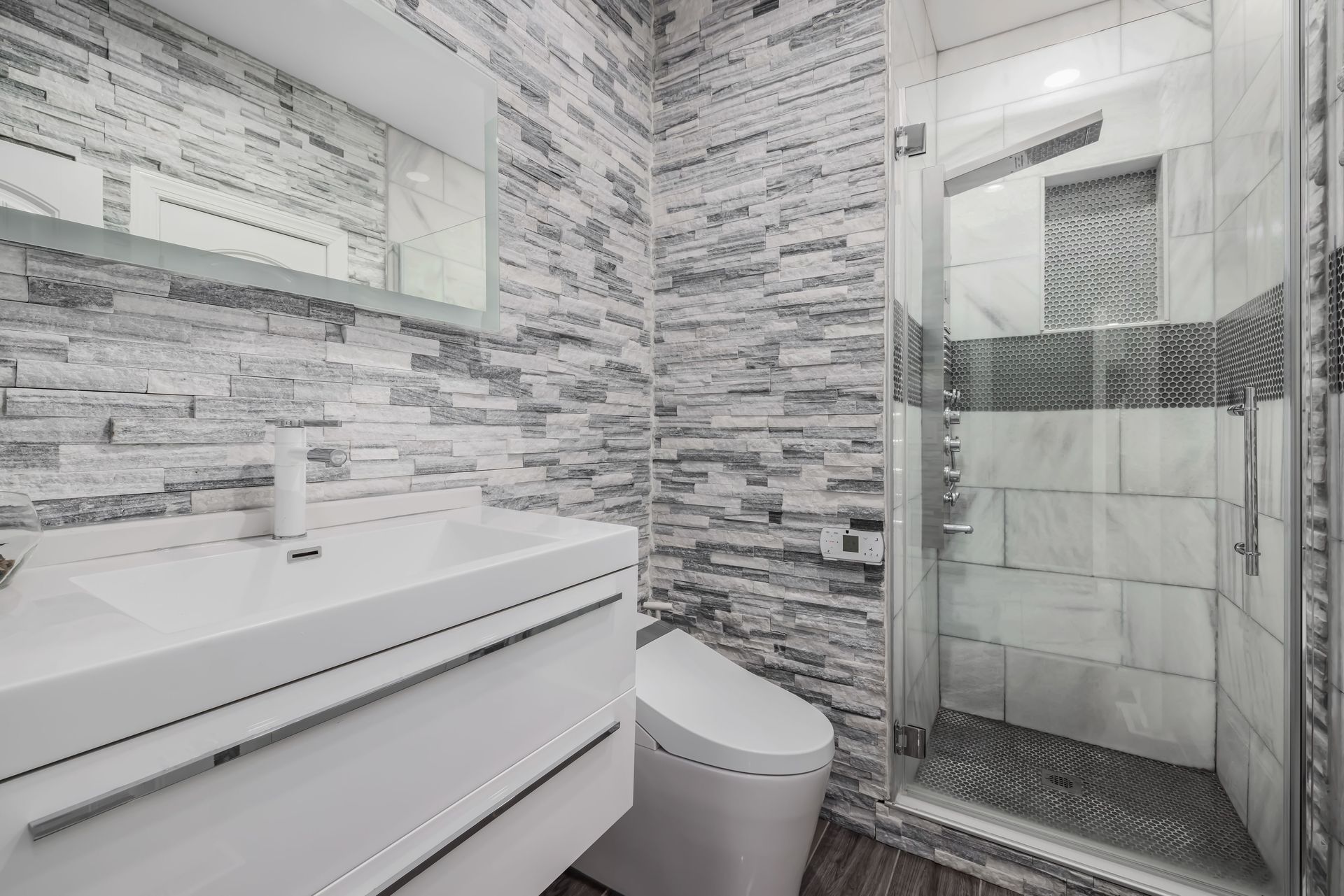 Modern bathroom with white vanity, toilet, and glass shower, accented by gray brick-pattern tile walls.