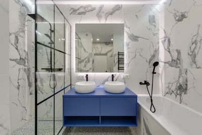 Modern bathroom with marble walls, blue double vanity, round sinks, mirror, glass shower, and bathtub.