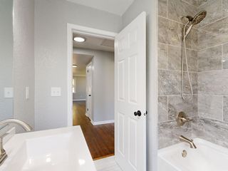 Bright bathroom with white tub, gray tile walls, and an open door to a hallway with hardwood floors