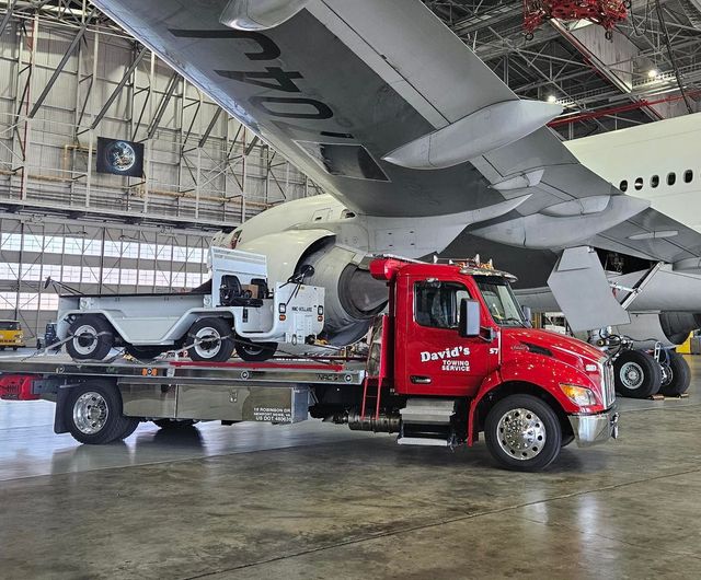 Red tow truck carrying a white jeep inside an airport hangar under a large airplane wing
