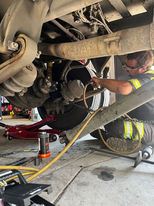 Mechanic working under a lifted vehicle in a garage, using tools near the rear wheel assembly.