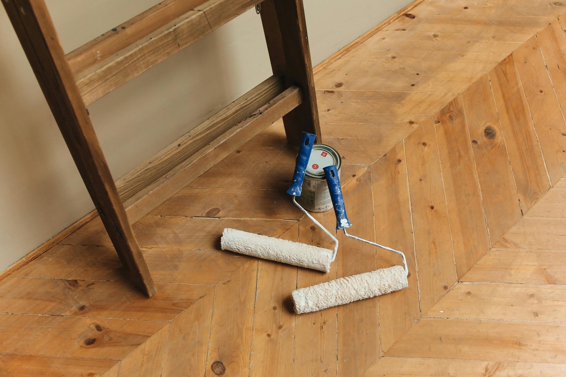 Wooden ladder leaning against wall, paint rollers on parquet floor.