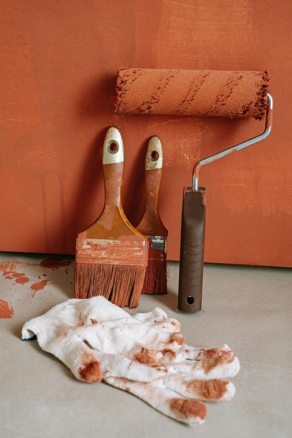 Paintbrushes, roller, and gloves stained with orange paint on a concrete surface next to a painted wall.