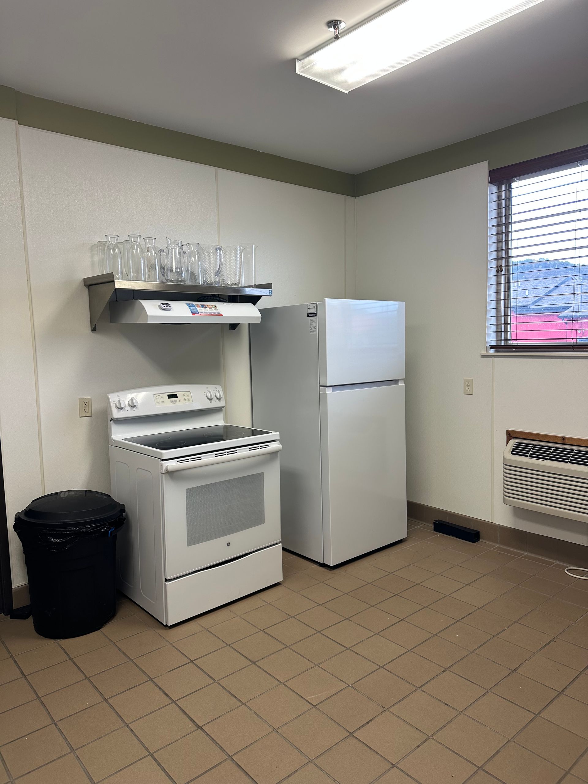 White kitchen appliances and a black trash can in a kitchen space with a window.