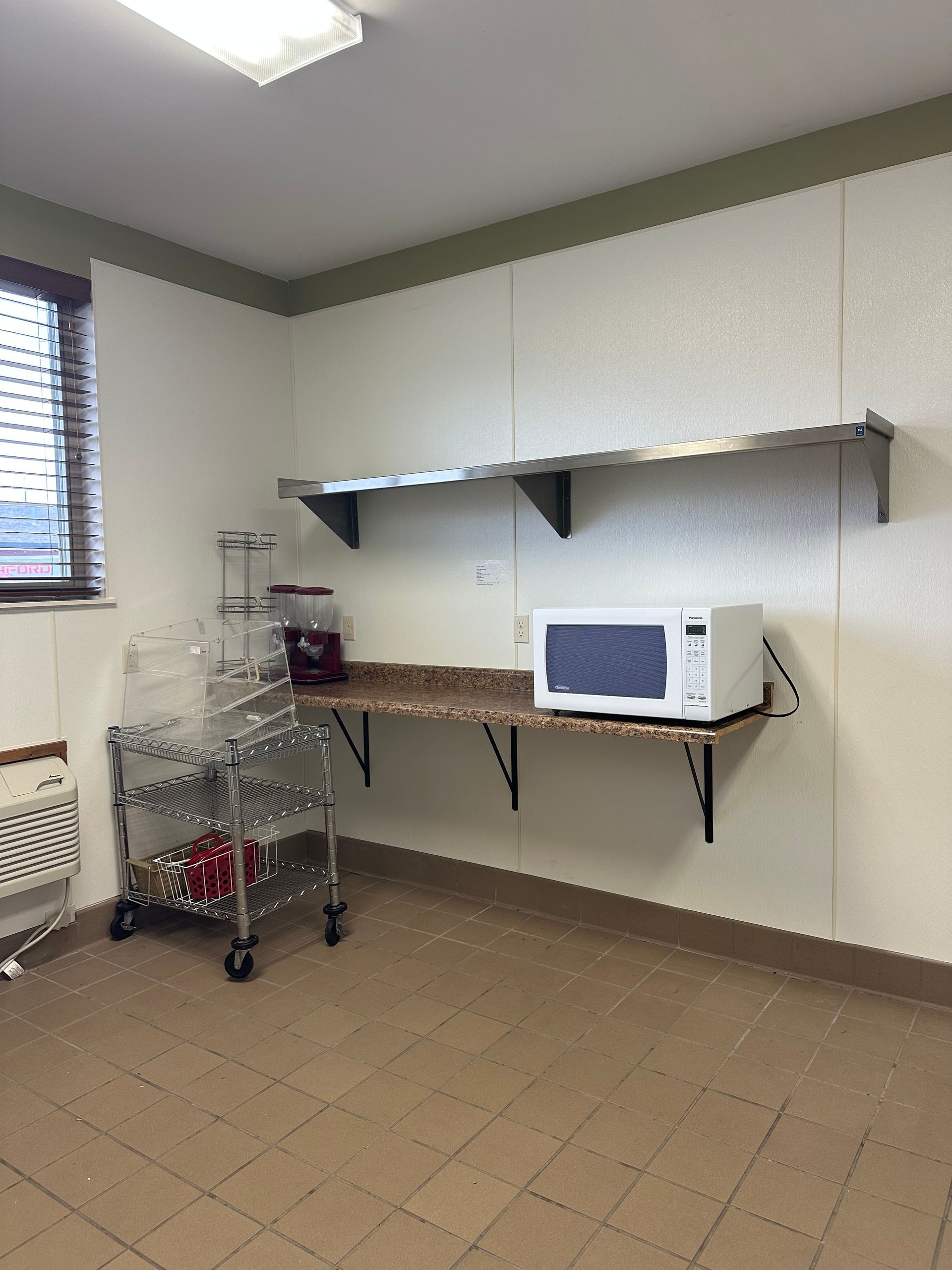 A small kitchen area with a microwave, shelving, and a rolling cart against a white wall and tan floor.