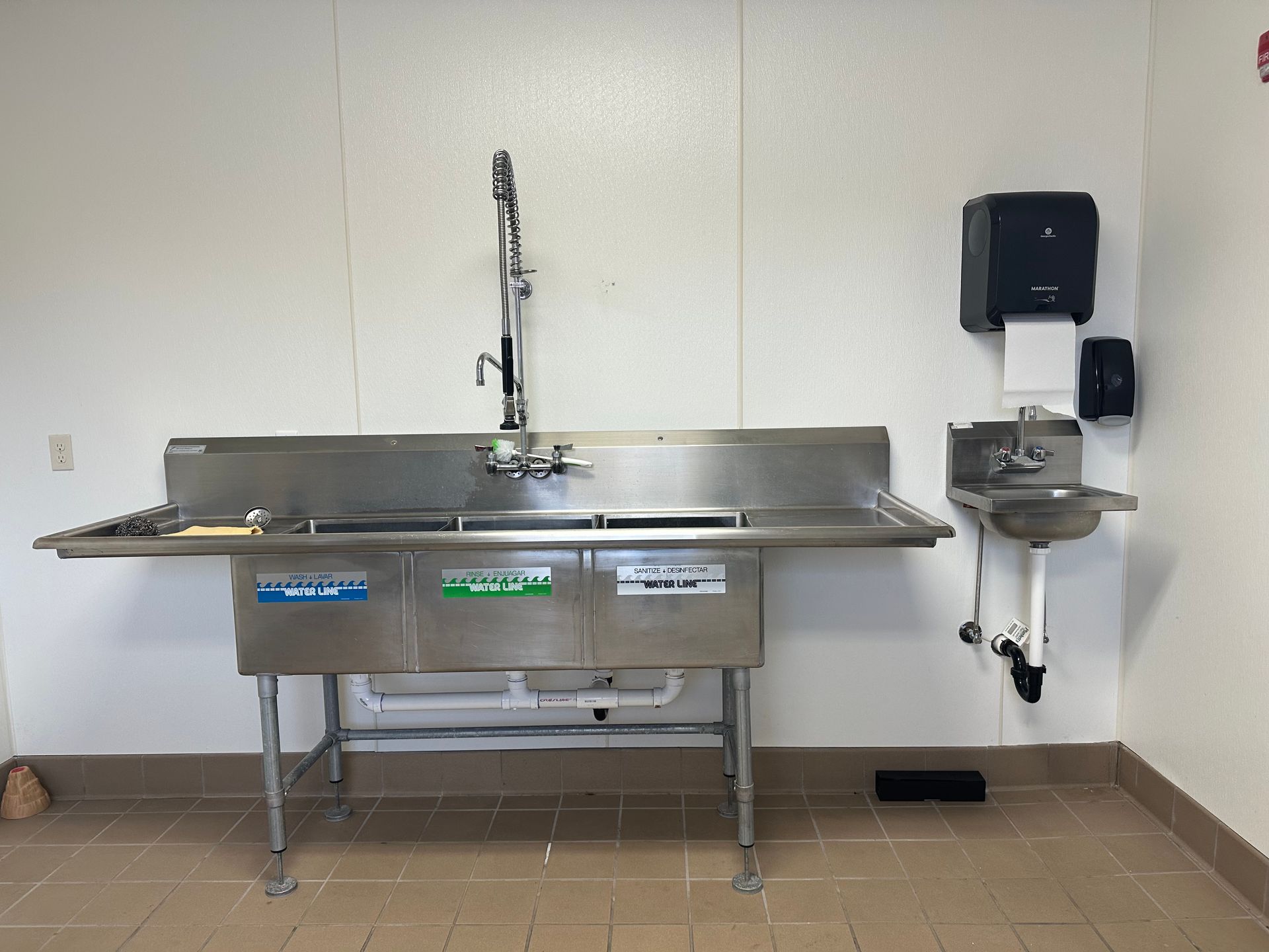 Stainless steel three-basin sink and small handwashing sink in a white-walled room.