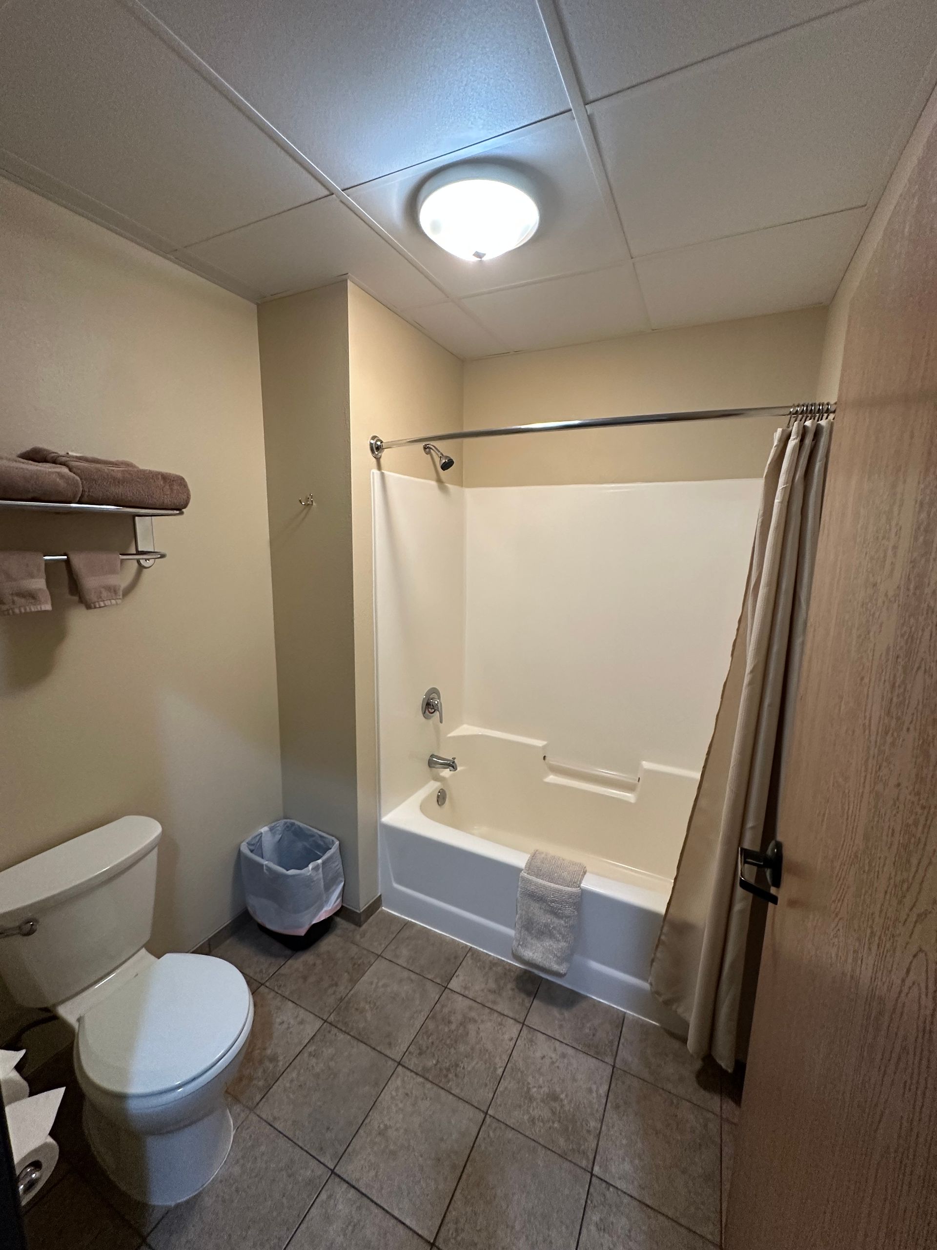 Bathroom with white toilet, tub, and beige walls, with tile floor, towel rack, and waste bin.