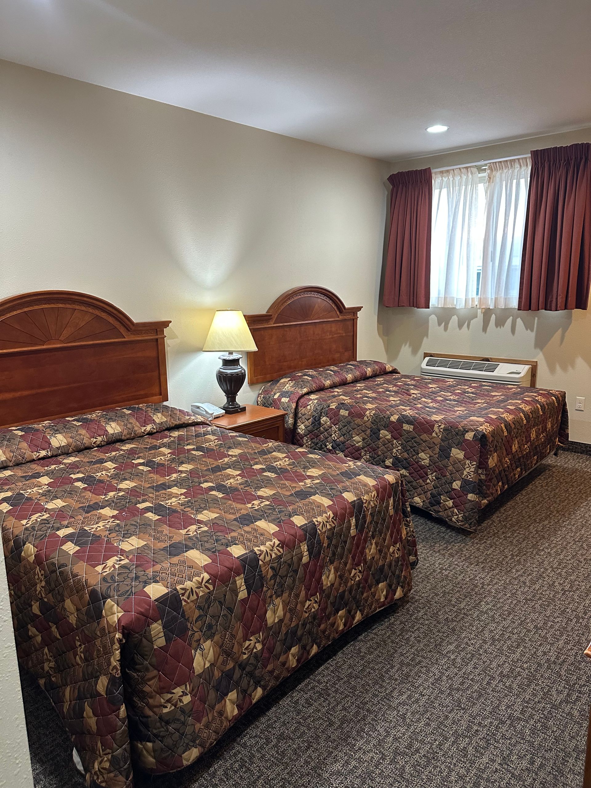 Two double beds in a motel room, featuring patterned comforters, a nightstand with a lamp, and a window with curtains.