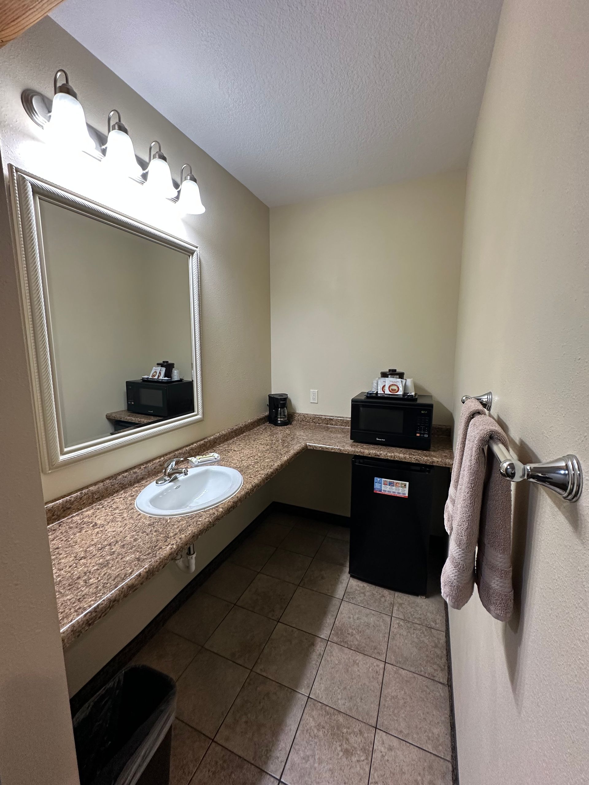 Bathroom with granite countertop, sink, mirror, microwave, mini-fridge, towels, and sconce lighting.