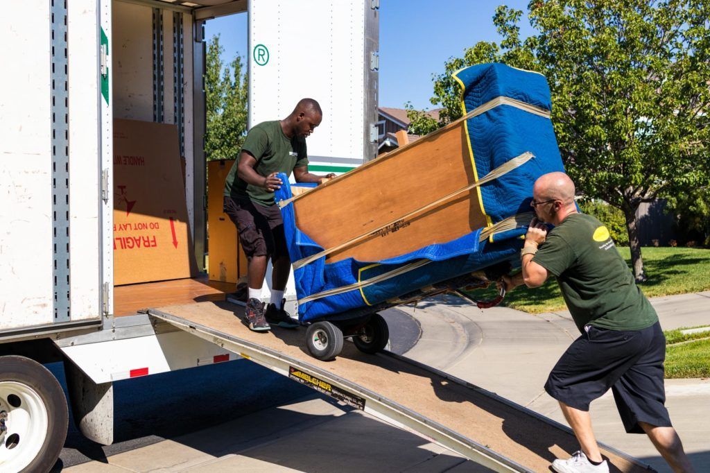 Two movers push a piano down a ramp from a truck.