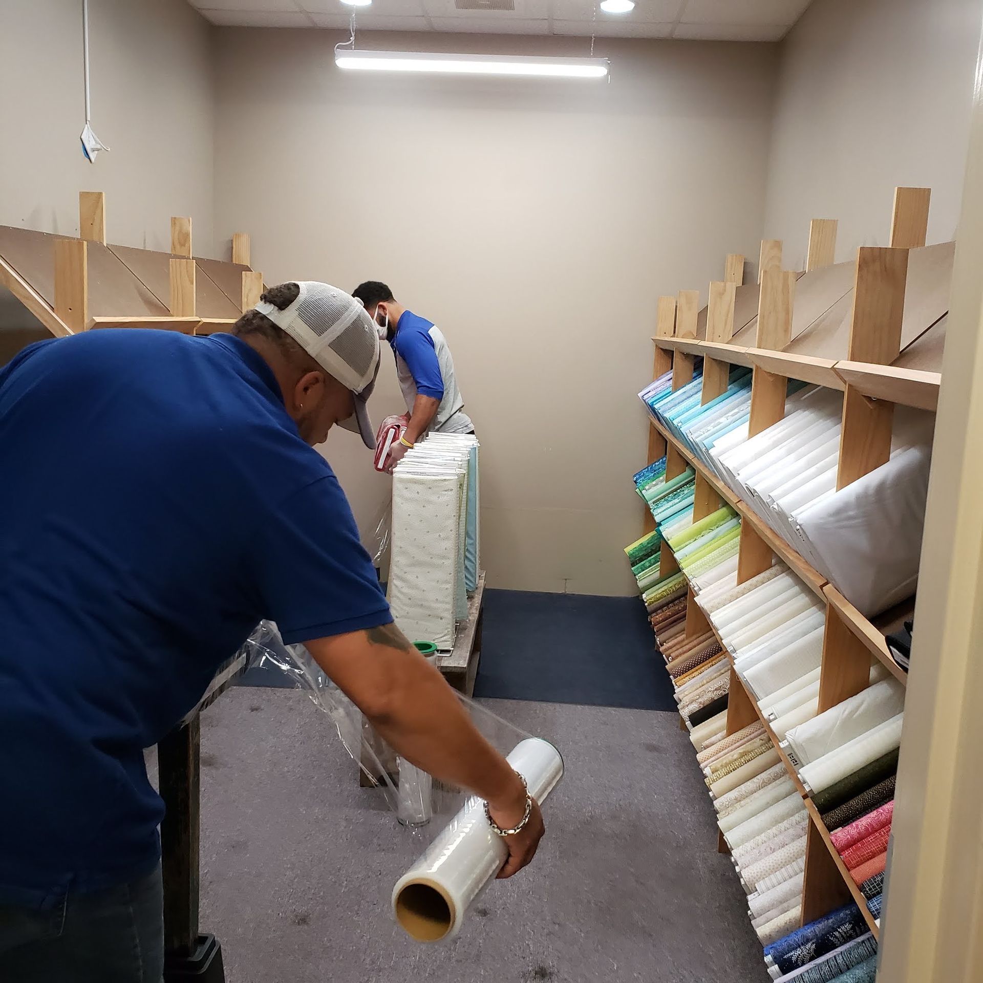 Two workers wrapping stacks of packaged goods in a small storage room with shelves.