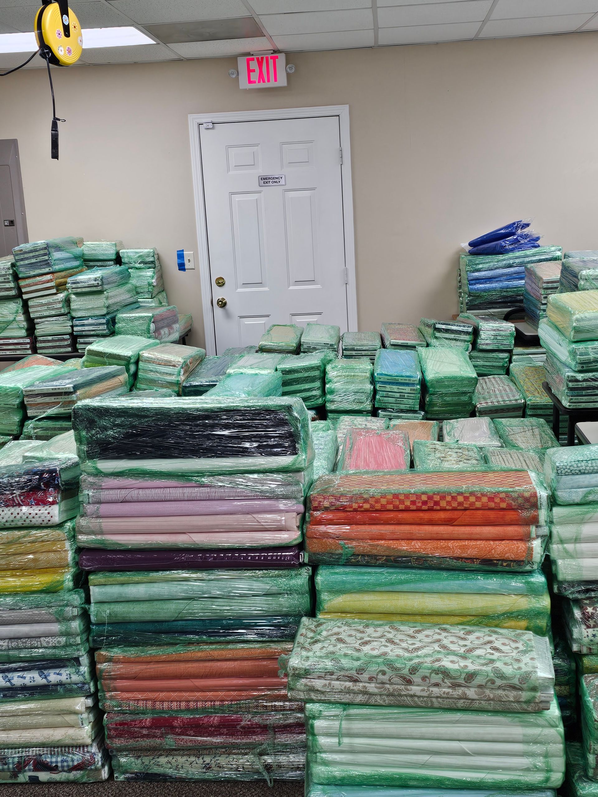 Stacks of plastic-wrapped folded mats or blankets piled in a room near a white door and EXIT sign.