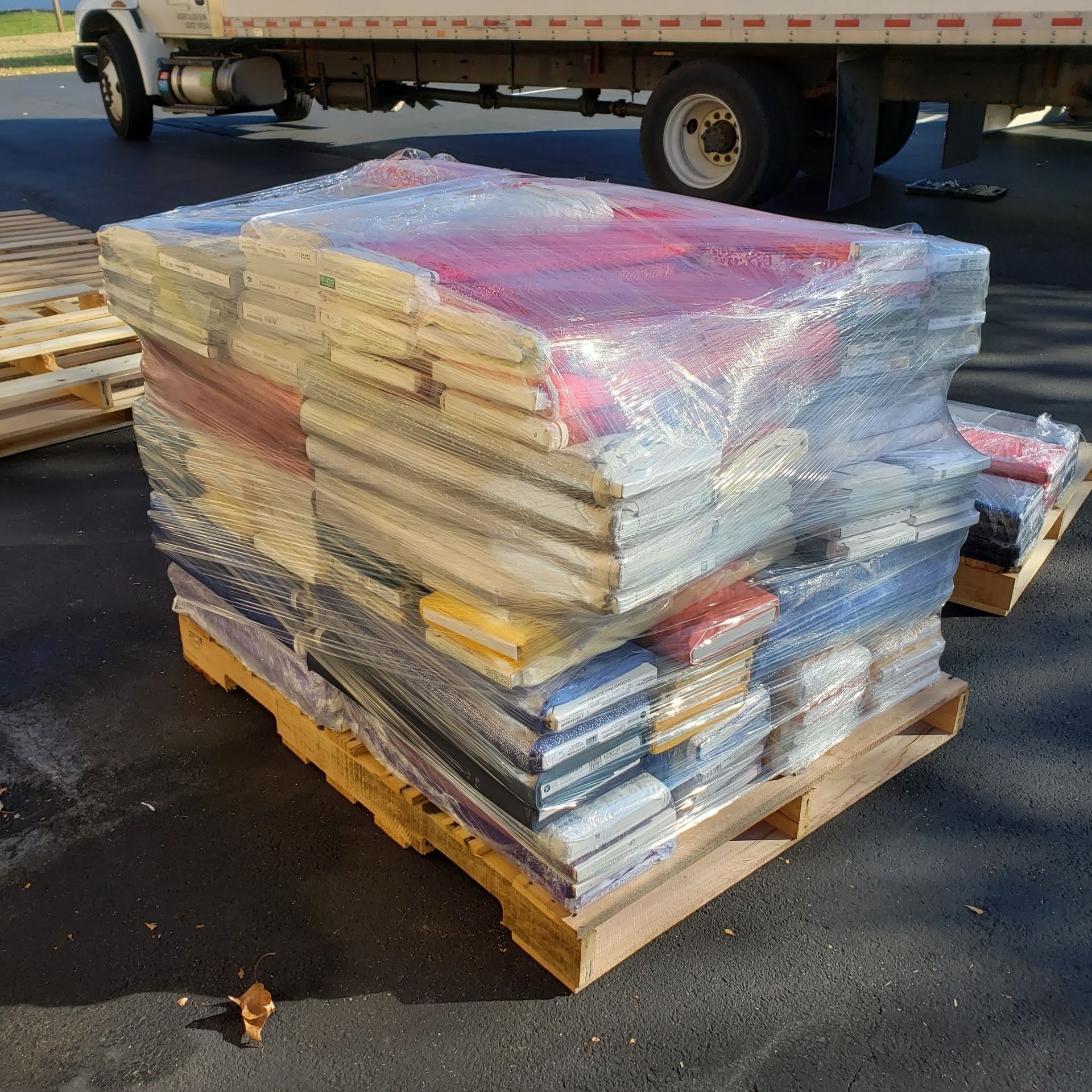 Shrink-wrapped stack of colorful flat packages on a wooden pallet in a parking lot, with a truck in the background